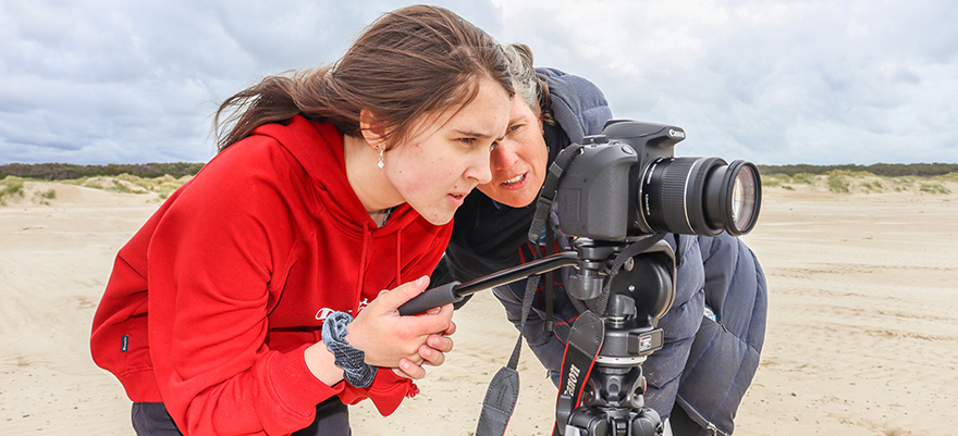 在圆形头部的海滩上站在相机后面的圆形头部和三脚架学习摄影的学生与导师一起学习摄影